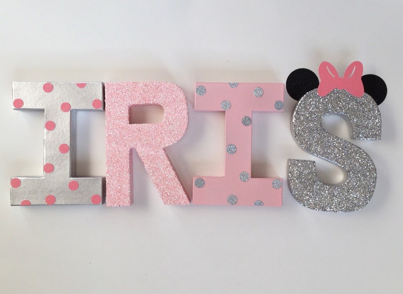 Op de afbeelding: Roze en zilveren glitterletters die de naam "IRIS" spellen met een zilveren glitterletter "S" met zwarte muizenoren en een roze strik.