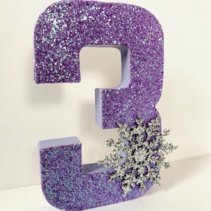 Pode incluir: N&uacute;mero tr&ecirc;s de glitter roxo com uma decora&ccedil;&atilde;o de floco de neve de glitter prata.