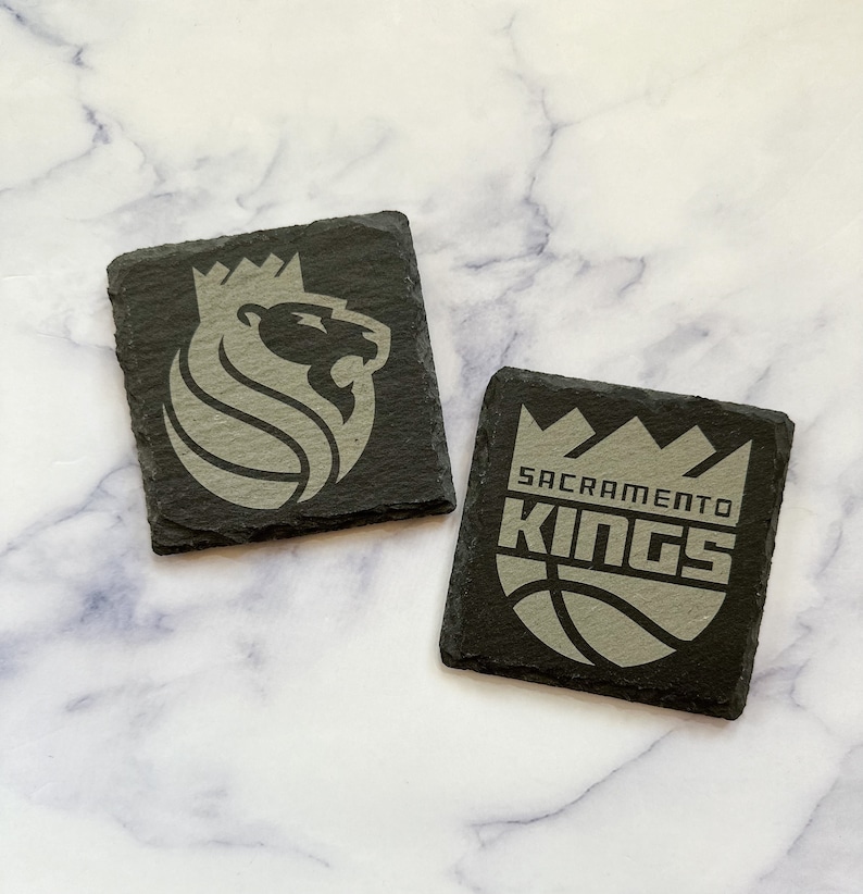May include: Two black slate coasters with the Sacramento Kings logo. One coaster features the team's lion mascot with a crown, and the other coaster features the team's name and a basketball.