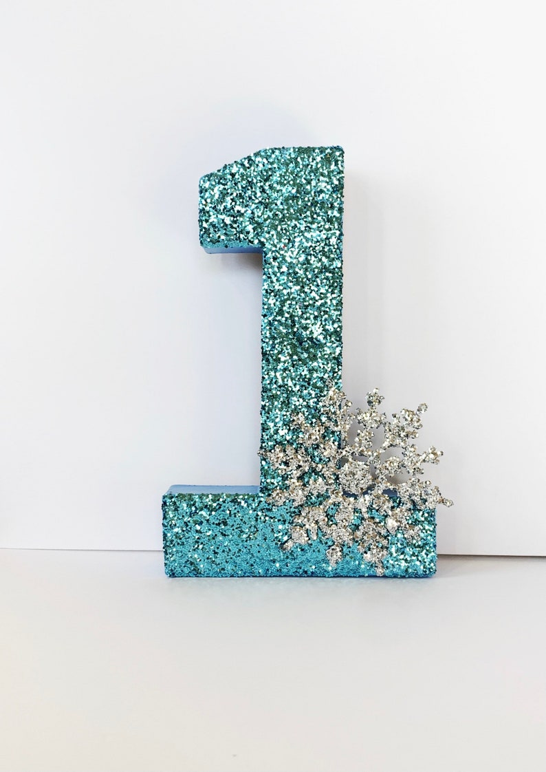 Pode incluir: Um n&uacute;mero um de glitter azul turquesa com uma decora&ccedil;&atilde;o de floco de neve de glitter prateado.