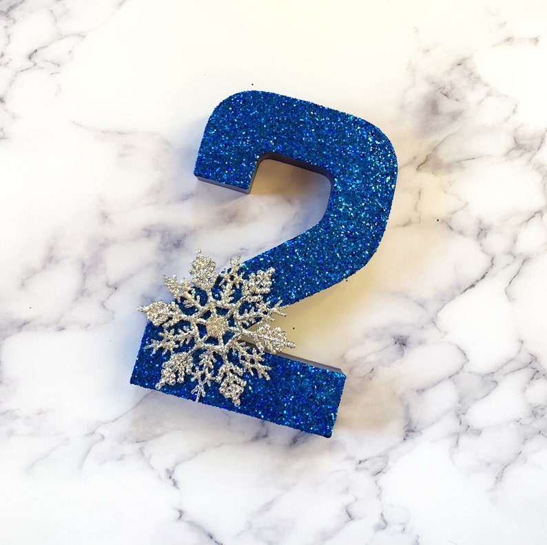 Pode incluir: Um n&uacute;mero dois de glitter azul com um floco de neve prateado no topo. O n&uacute;mero est&aacute; sobre um fundo de m&aacute;rmore branco.