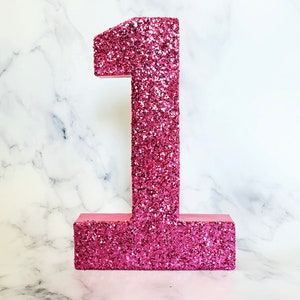 Pode incluir: N&uacute;mero um de glitter rosa. O n&uacute;mero &eacute; feito de papel&atilde;o e coberto de glitter rosa. &Eacute; um n&uacute;mero decorativo para uma festa ou celebra&ccedil;&atilde;o.