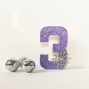 Pode incluir: N&uacute;mero tr&ecirc;s de purpurina roxa com uma decora&ccedil;&atilde;o de floco de neve prateado. Tr&ecirc;s bolas de discoteca prateadas est&atilde;o em primeiro plano.