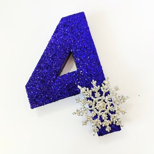 Pode incluir: Um n&uacute;mero quatro de glitter azul com uma decora&ccedil;&atilde;o de floco de neve de glitter prateado.
