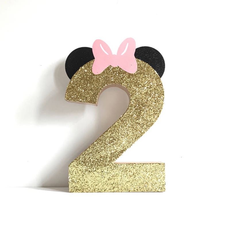 Pu&ograve; includere: Numero due in glitter dorato con orecchie da topo nere e un fiocco rosa in cima.