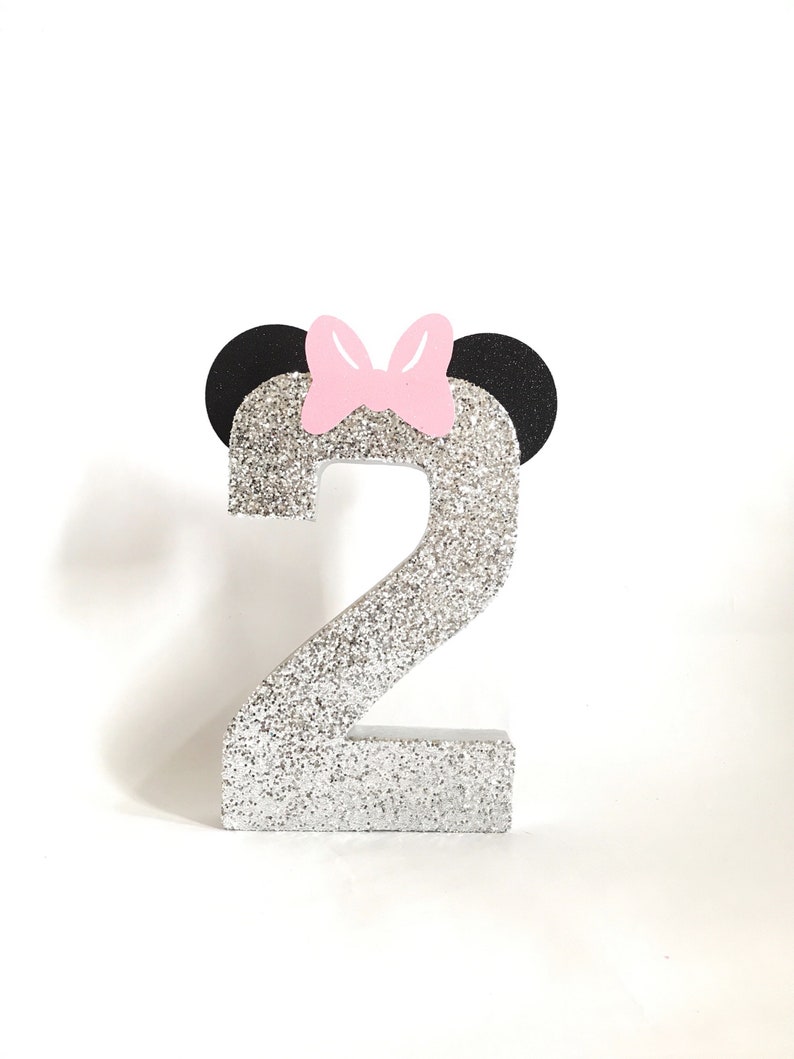 Pu&ograve; includere: Numero due di glitter argento con orecchie nere e un fiocco rosa in cima.