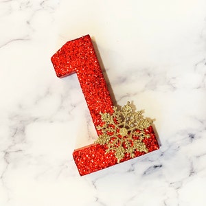 Pode incluir: N&uacute;mero um vermelho com glitter com um floco de neve dourado com glitter sobre um fundo de m&aacute;rmore branco.