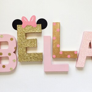 Op de afbeelding: Roze en gouden glitterletters die de naam "BELA" spellen met een roze strik en zwarte oren op de "E".