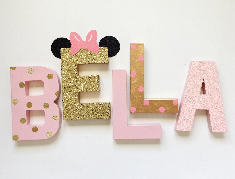 Op de afbeelding: Roze en gouden glitterletters die de naam "BELA" spellen met een roze strik en zwarte oren op de "E".