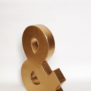 May include: A gold ampersand symbol made of cardboard. The symbol is standing upright on a white surface.