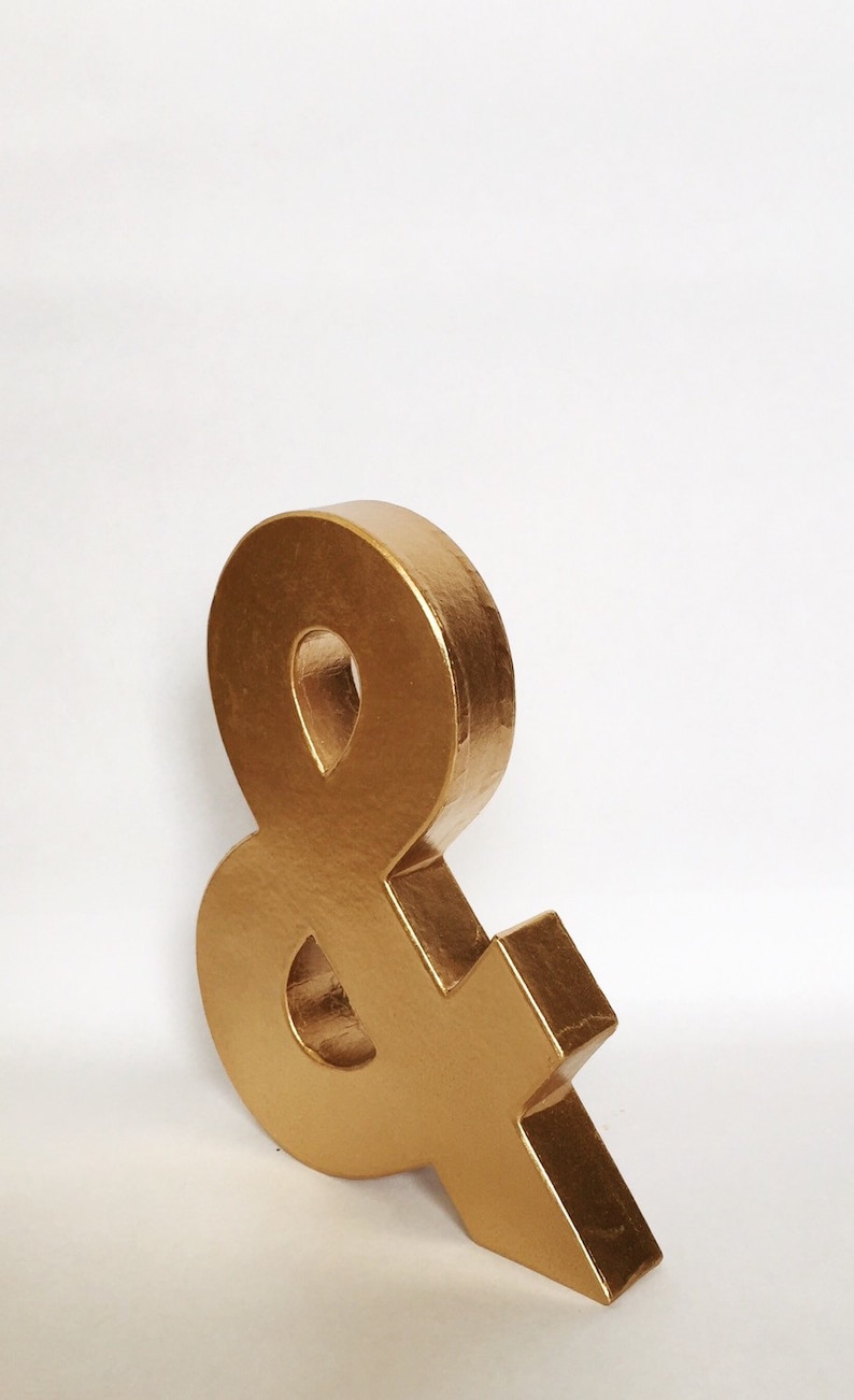 May include: A gold ampersand symbol made of cardboard. The symbol is standing upright on a white surface.
