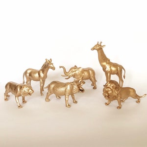 May include: A set of six gold-painted plastic animal figurines, including a zebra, a hippopotamus, a giraffe, a lion, and an elephant. The figurines are all standing on a white surface.