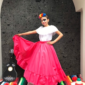 May include: A young person in a traditional Mexican costume, featuring a white blouse with lace trim and a vibrant pink skirt. A colorful flower crown adorns their head, and a decorative guitar rests nearby. The scene is set against a dark textured wall.
