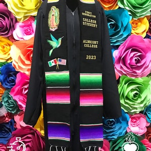 May include: Black graduation stole with colorful stripes and embroidered details. The stole features a hummingbird, the Virgin Mary, and the text "First Generation College Student", "Albright College", and "2023". The stole is displayed against a background of colorful paper flowers.