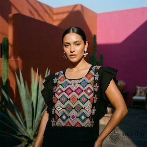 Op de afbeelding: Een zwarte blouse met ruches aan de mouwen en een centraal paneel versierd met een levendig, veelkleurig geometrisch ruitpatroon. Het ontwerp bevat kleine gestileerde figuren aan de zijkanten van het geborduurde borstgedeelte.
