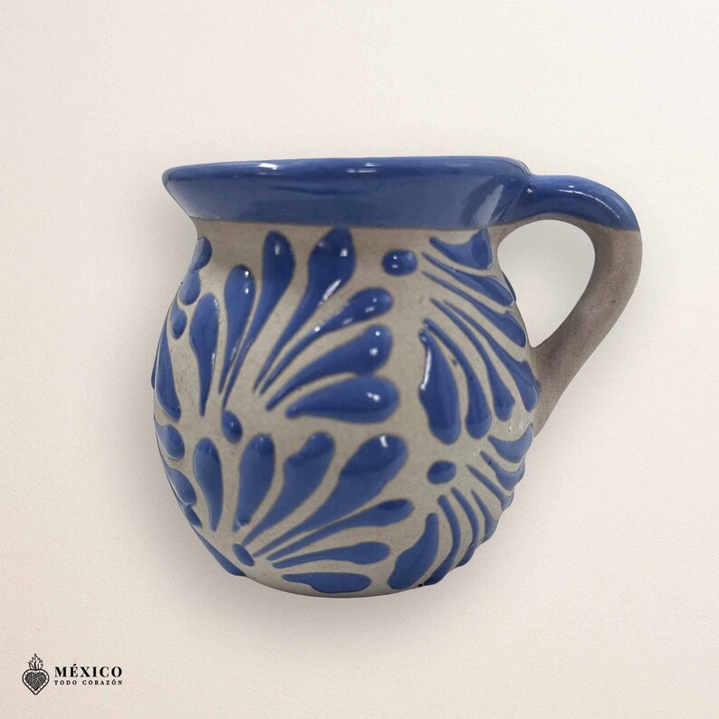 Peut inclure: Une tasse en c&eacute;ramique artisanale avec un bord et une anse bleus. La tasse pr&eacute;sente une base de couleur cr&egrave;me orn&eacute;e d'un motif floral bleu r&eacute;p&eacute;titif. La poign&eacute;e est de couleur grise. La tasse a une forme et un design traditionnels.