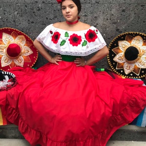 May include: A white off-the-shoulder blouse with red poppy embroidery and lace trim, paired with a long red skirt featuring a green waistband. The outfit includes a red fabric flower headpiece and a matching red choker, with two large sombreros nearby.