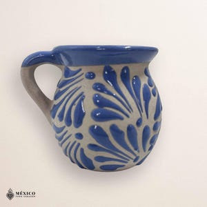 Peut inclure: Une tasse en c&eacute;ramique avec un bord et une anse bleus, orn&eacute;e d'un corps gris d&eacute;cor&eacute; de motifs floraux bleus. La tasse a une anse incurv&eacute;e et un bord l&eacute;g&egrave;rement &eacute;vas&eacute;. Le design rappelle la poterie mexicaine traditionnelle.