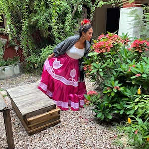 May include: A woman wearing a pink skirt with white lace trim and floral embroidery, a white top, and a black and white patterned shawl. She is standing in a garden and smelling a red flower.