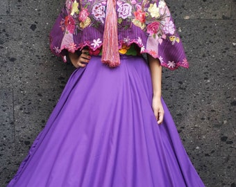 May include: A woman wearing a purple skirt with a ruffled hem and a purple embroidered shawl with floral designs. She is wearing a purple headband and earrings.