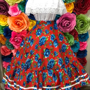 Puede incluir: Una falda roja con estampado de rosas azules y ribete blanco. La falda es amplia y tiene un aspecto fruncido. Es una falda tradicional mexicana.