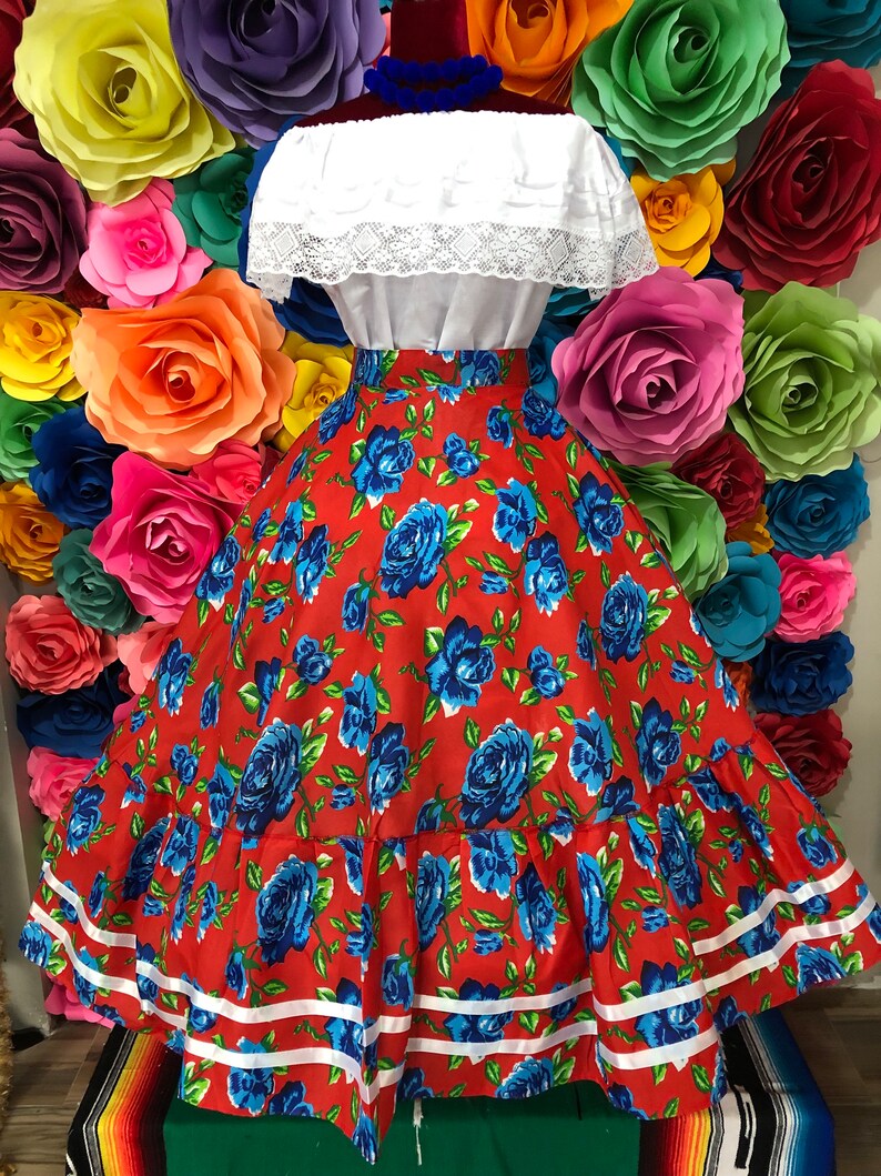 Puede incluir: Una falda roja con estampado de rosas azules y ribete blanco. La falda es amplia y tiene un aspecto fruncido. Es una falda tradicional mexicana.