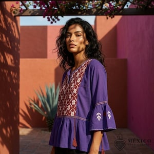 Op de afbeelding: Een paarse blouse met een V-hals en een donkerrood en wit geometrisch geborduurd paneel op de borst. Het kledingstuk heeft driekwart klokmouwen met witte bladerenborduurwerk en een geplooide peplum-taille met twee kwastjes aan de voorzijde.