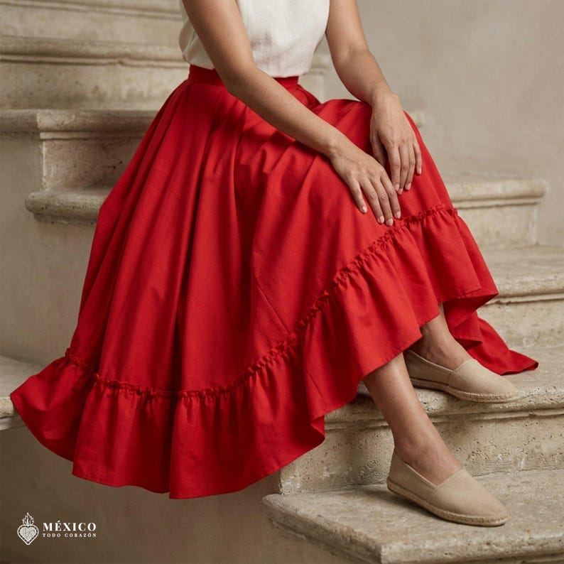 May include: A vibrant red midi skirt featuring a high-waisted design with a gathered, ruffled hemline. The skirt is paired with a simple white sleeveless top and beige slip-on espadrilles. The fabric appears smooth and solid in color.