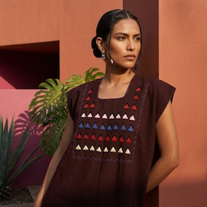 Puede incluir: Una túnica sin mangas de color marrón oscuro con escote cuadrado y un panel central de motivos geométricos triangulares bordados en rojo, rosa, azul y crema. El bordado está enmarcado por un fino borde azul cosido en forma ondulada.