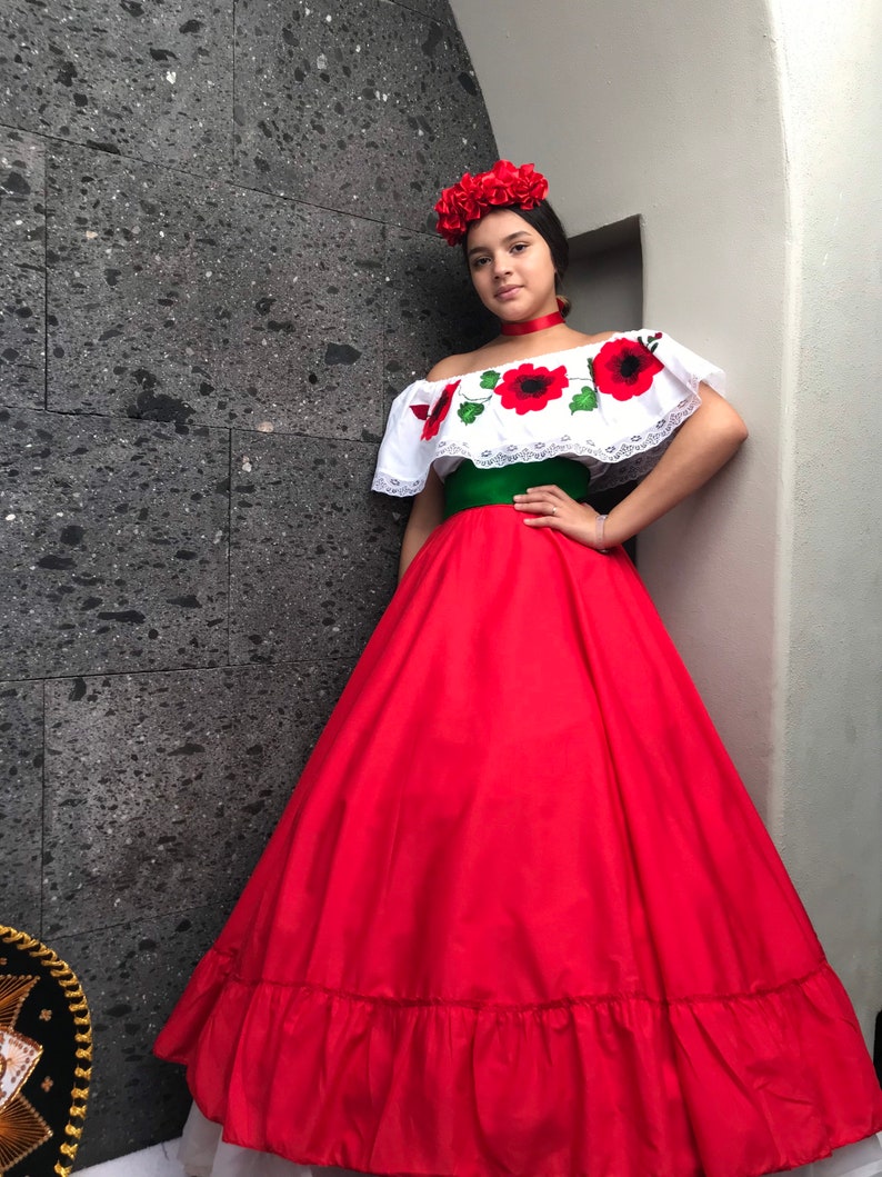 May include: A floor-length red skirt with a ruffled hem, paired with an off-the-shoulder white blouse featuring red and black floral embroidery and lace trim. A wide green sash sits at the waist, and a red fabric flower crown is worn on the head.