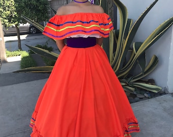 May include: A young person wearing a traditional Mexican dress with an orange skirt, a white top with colorful stripes, and a purple sash. The dress has a ruffled hem with colorful ribbons. The person is wearing a flower crown with purple flowers.