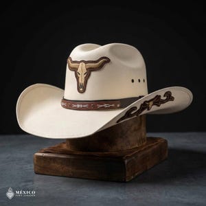 Op de afbeelding: Een crèmekleurige cowboyhoed met een bruin geborduurd stierenschedel-embleem op de voorkant van de kroon. De hoed heeft een bruine leren band met witte stiksels en metalen studs, plus decoratief bruin borduurwerk op de brede, gebogen rand.