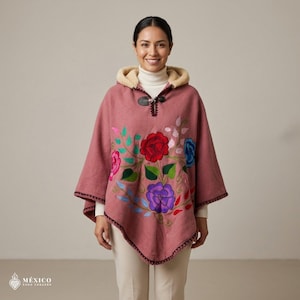 May include: A dusty rose hooded poncho featuring vibrant floral embroidery in red, blue, and purple with green leaves. The garment includes a cream-colored fleece-lined hood, a single toggle closure at the neckline, and dark decorative stitching along the hem.