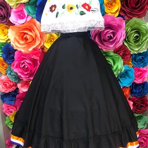 May include: A traditional Mexican dress, featuring a white embroidered top with floral designs and a long black skirt. The skirt has a ruffled hem with orange, white, and purple ribbon trim. The dress is displayed against a backdrop of colorful paper flowers.