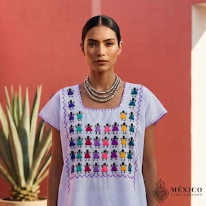 Puede incluir: Una túnica de manga corta en color azul claro con escote cuadrado y un patrón bordado multicolor de figuras estilizadas en morado, rosa, amarillo y turquesa. La prenda incluye un collar plateado de varias capas y costuras moradas.