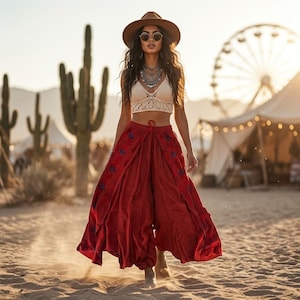 May include: A cream-colored crochet crop top with a V-neckline and a long, flowing dark red skirt featuring blue floral embroidery along the front panels. The outfit is paired with a wide-brimmed brown hat and round sunglasses.