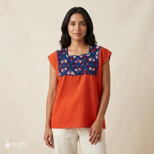 Puede incluir: Una blusa naranja de manga corta con cuello cuadrado. El cuello presenta un diseño bordado en azul con detalles rojos y morados. La imagen incluye la palabra "MÉXICO" y un símbolo de corazón.