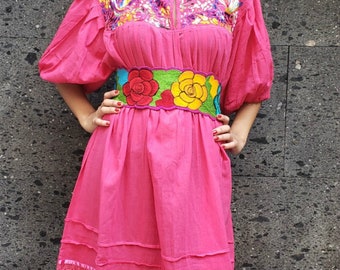 May include: A pink embroidered dress with a floral embroidered belt. The dress has a round neckline and puffed sleeves. The dress has a lace trim at the bottom.