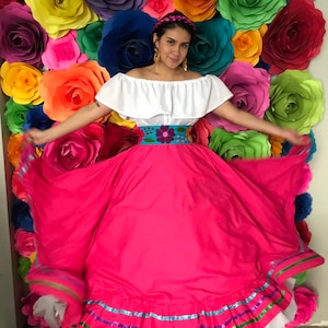 May include: A woman wearing a white off-the-shoulder blouse with a ruffled neckline and a pink skirt with colorful embroidered trim. She is standing in front of a wall decorated with large paper flowers in various colors.