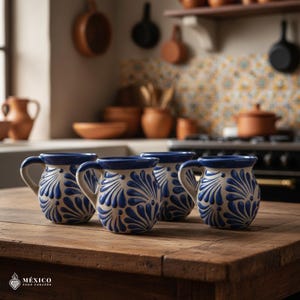 Peut inclure: Quatre tasses en c&eacute;ramique avec une base cr&egrave;me et un motif floral bleu sont pos&eacute;es sur une table en bois. Les tasses ont un bord et une anse bleus. L'arri&egrave;re-plan montre une cuisine avec des pots et des casseroles en terre cuite.