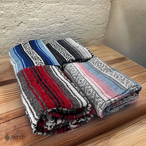 May include: Four folded Mexican blankets in various colors, including blue, black, red, and pink. Each blanket features a striped pattern with a woven design. The blankets are stacked on a wooden surface.