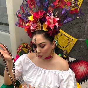 Puede incluir: Una mujer con una blusa blanca de hombros descubiertos y una falda amarilla. Lleva una gran diadema floral con un abanico de encaje negro. Sostiene un rosario marrón en sus manos.