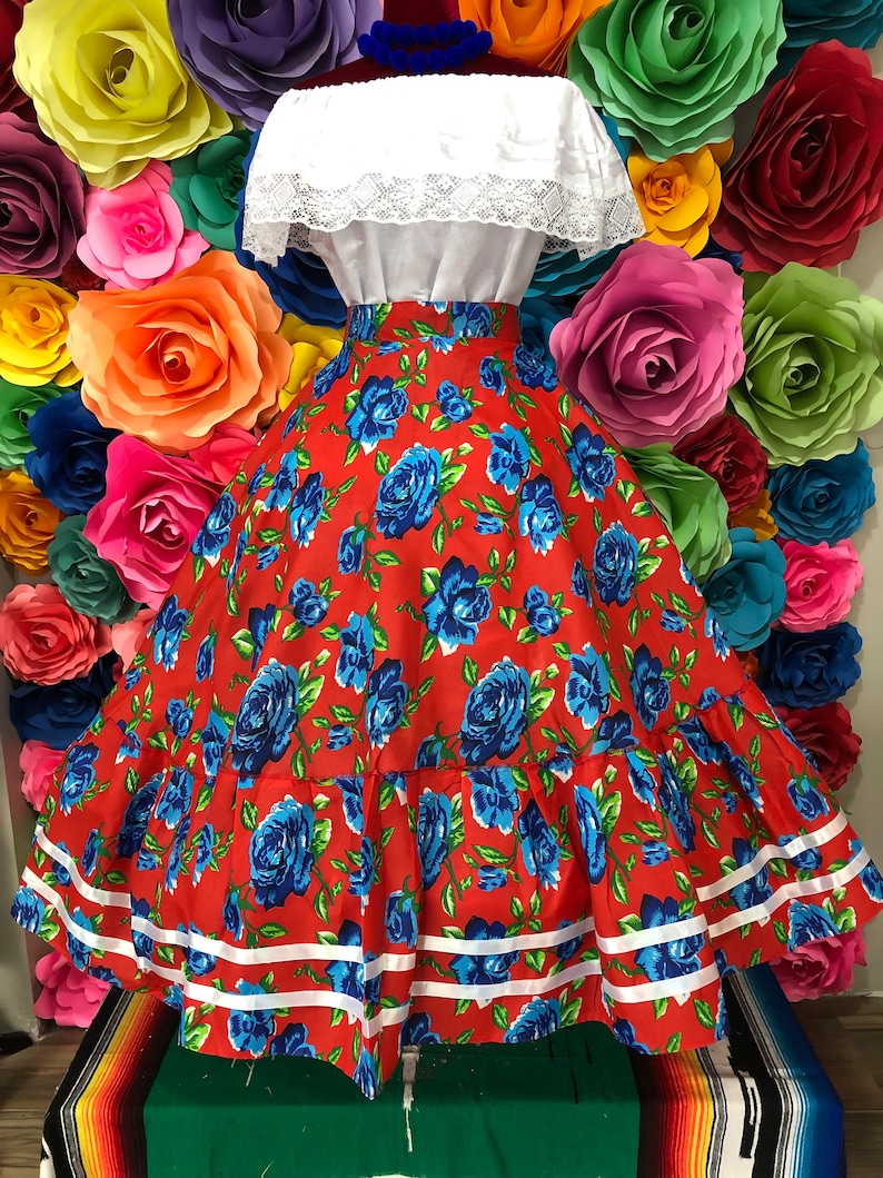 Puede incluir: Una falda roja con estampado floral azul y ribete blanco. La falda es amplia y fluida, y tiene una cintura el&aacute;stica. La falda est&aacute; siendo usada por un maniqu&iacute; contra un fondo de flores de papel de colores.