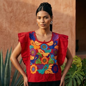 Puede incluir: Una blusa roja sin mangas con escote cuadrado y detalles plisados en los hombros. La parte delantera está decorada con bordados vibrantes y coloridos de flores y pájaros en tonos amarillo, azul, verde, naranja y morado.