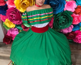 May include: A woman wearing a traditional Mexican dress, featuring a green skirt and off-the-shoulder top with colorful trim. She wears a red choker and a floral headpiece, posing in front of a backdrop of large, colorful paper roses.
