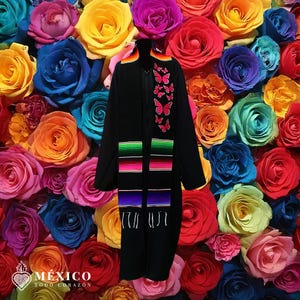 May include: A black graduation stole with colorful embroidered details. The stole features a vibrant striped design and pink butterfly accents. The background is a wall of colorful roses. The stole has the text "MEXICO TODO CORAZÓN" at the bottom.