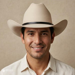 Puede incluir: Un sombrero de vaquero de fieltro beige con una copa alta con pliegue central y un ala ancha y curvada. El sombrero está acentuado por una fina cinta negra texturizada que rodea la base de la copa, creando un contraste con el color beige claro.
