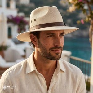 Peut inclure: Un chapeau fedora de couleur crème avec une calotte en maille tissée texturée et un large bord. Le chapeau présente une bande en cuir marron foncé entourant la base de la calotte et deux petits œillets de ventilation métalliques sur chaque côté.