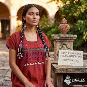 Puede incluir: Una blusa color óxido de manga corta con escote cuadrado y filas de figuras bordadas en rosa, turquesa, morado y blanco. Un letrero muestra las dimensiones: 53,3 cm de ancho, 22,9 cm de sisa y 61 cm de largo, para tallas S-L.