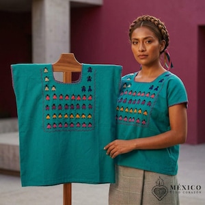 Puede incluir: Una blusa de manga corta color verde azulado con escote cuadrado y filas bordadas de pequeñas figuras en rosa, naranja, morado y beige. La prenda se muestra en una percha de madera y la lleva puesta una persona con el cabello trenzado.